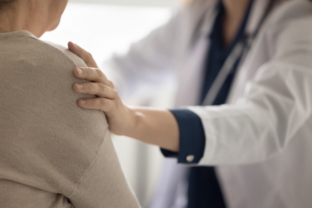 Close-up cropped shot of a geriatrician doctor's hand gently touching the shoulder of an elderly patient woman, conveying empathy, comfort, and psychological support while discussing a serious illness diagnosis.
