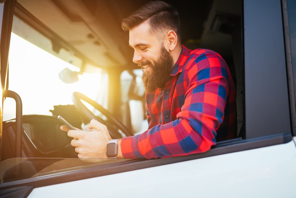 A youthful and attractive man with a beard is seen using his smartphone while seated in his truck.