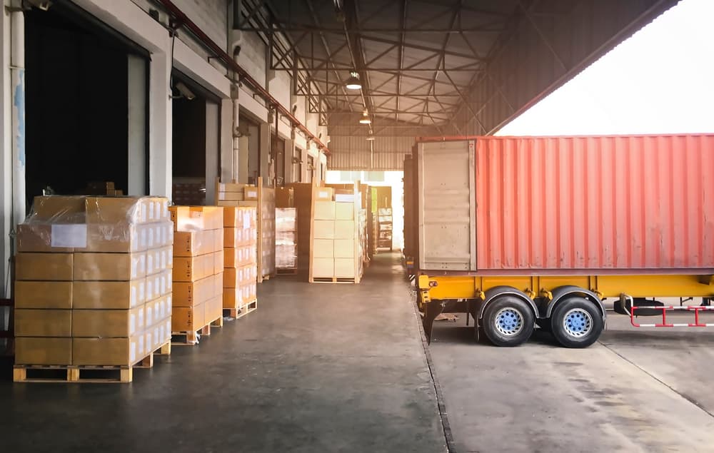 Un camión con remolque está aparcado en un muelle de almacén, cargando cajas de paquetes en un contenedor de carga. Esta actividad representa el proceso esencial del envío de carga dentro de la cadena de suministro.