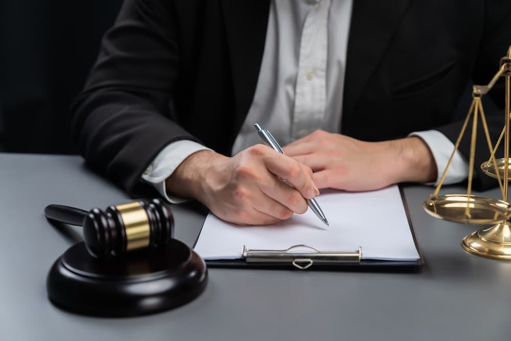 Symbols of justice, including a gavel and balance scales, in focus with a blurred background of a lawyer or judge signing a legal document at a desk, representing integrity, fairness, and equality in the legal system.