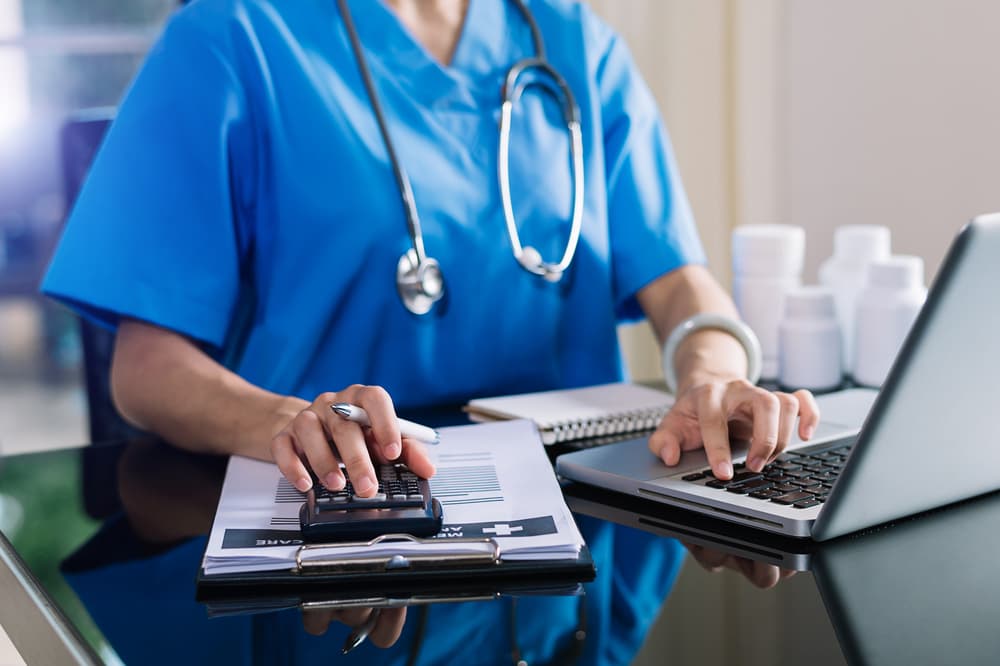 Concepto de gastos y honorarios sanitarios. Un médico experto utiliza una calculadora y un smartphone, junto con una tableta, para gestionar los gastos médicos en un hospital.