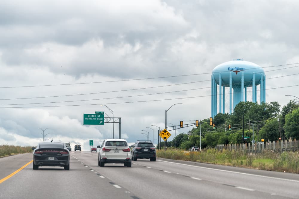 Autopista interestatal en Fort Worth, Texas, con numerosos coches circulando y un cartel sobre un depósito de agua al fondo.