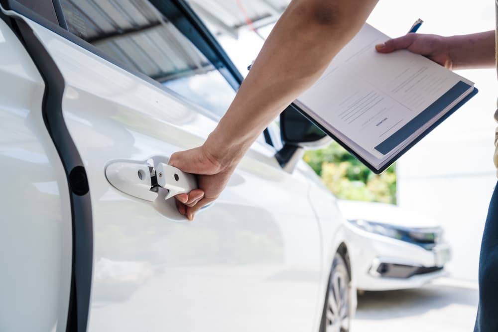 A person inspecting a white car's door handle while holding a clipboard with documents.