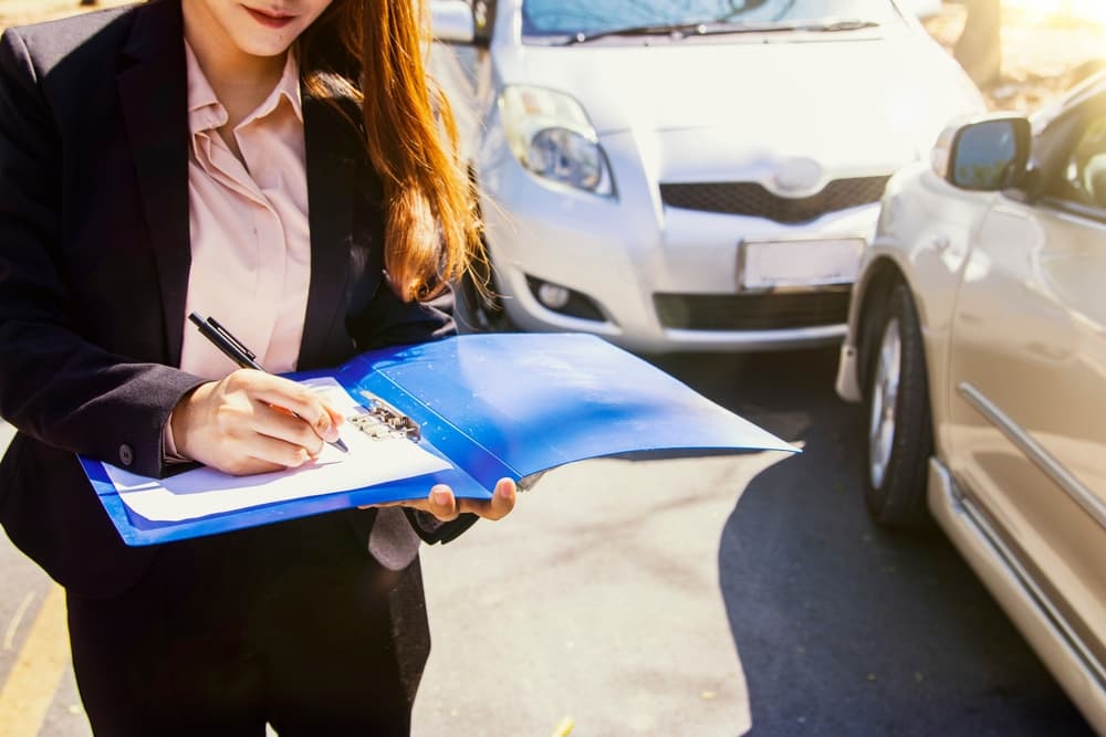 Mujer aseguradora en el lugar de un accidente de coche, tomando notas y recopilando pruebas para reclamaciones de seguros y disputas por indemnizaciones entre las partes.