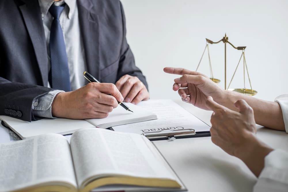 Car accident lawyer and woman discuss documents at a desk, taking notes. Scales of justice and book visible, indicating a legal setting.