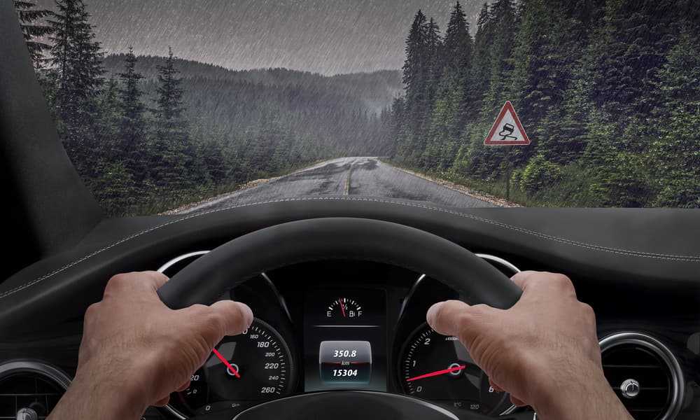 Driver's view in rainy weather with hands on the wheel, passing a slippery road sign, rain splashing on windshield.