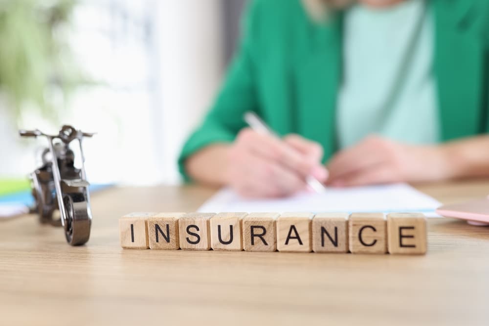 Vehicle and health insurance concept with motorcycle model and wooden blocks spelling 'insurance' on table, manager holding document and pen in the background.