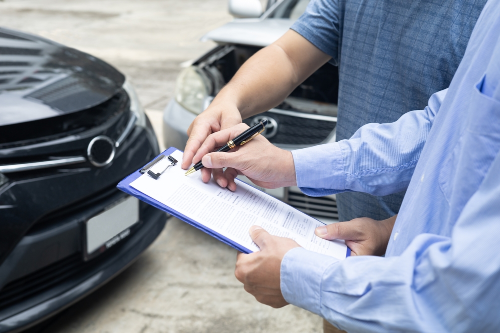 Un funcionario de seguros escribe en un portapapeles mientras un agente de seguros examina un coche negro, revisando las pólizas de seguros de accidentes de coche.