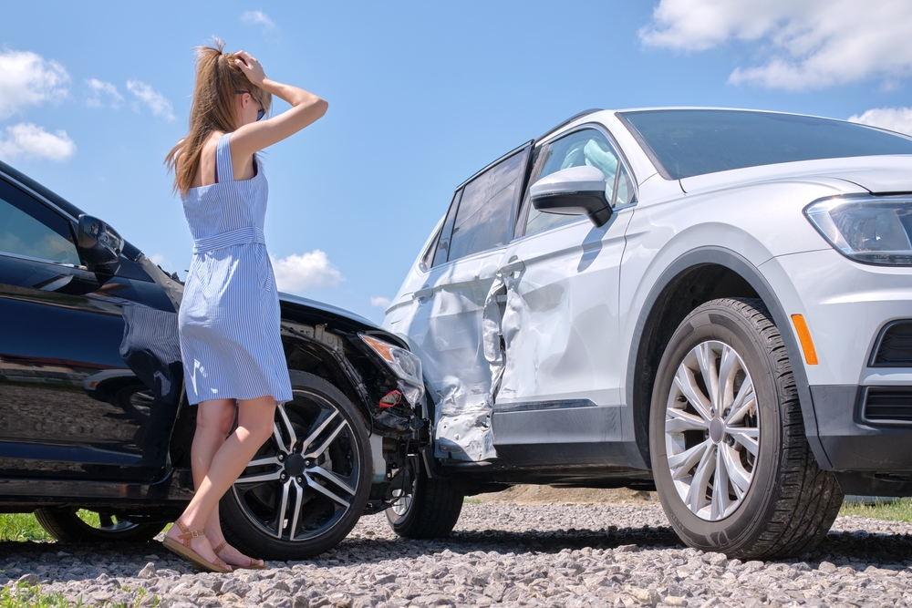 Triste joven conductora de pie cerca de su coche destrozado con cara de conmoción tras un accidente de giro a la izquierda en una carretera muy transitada.